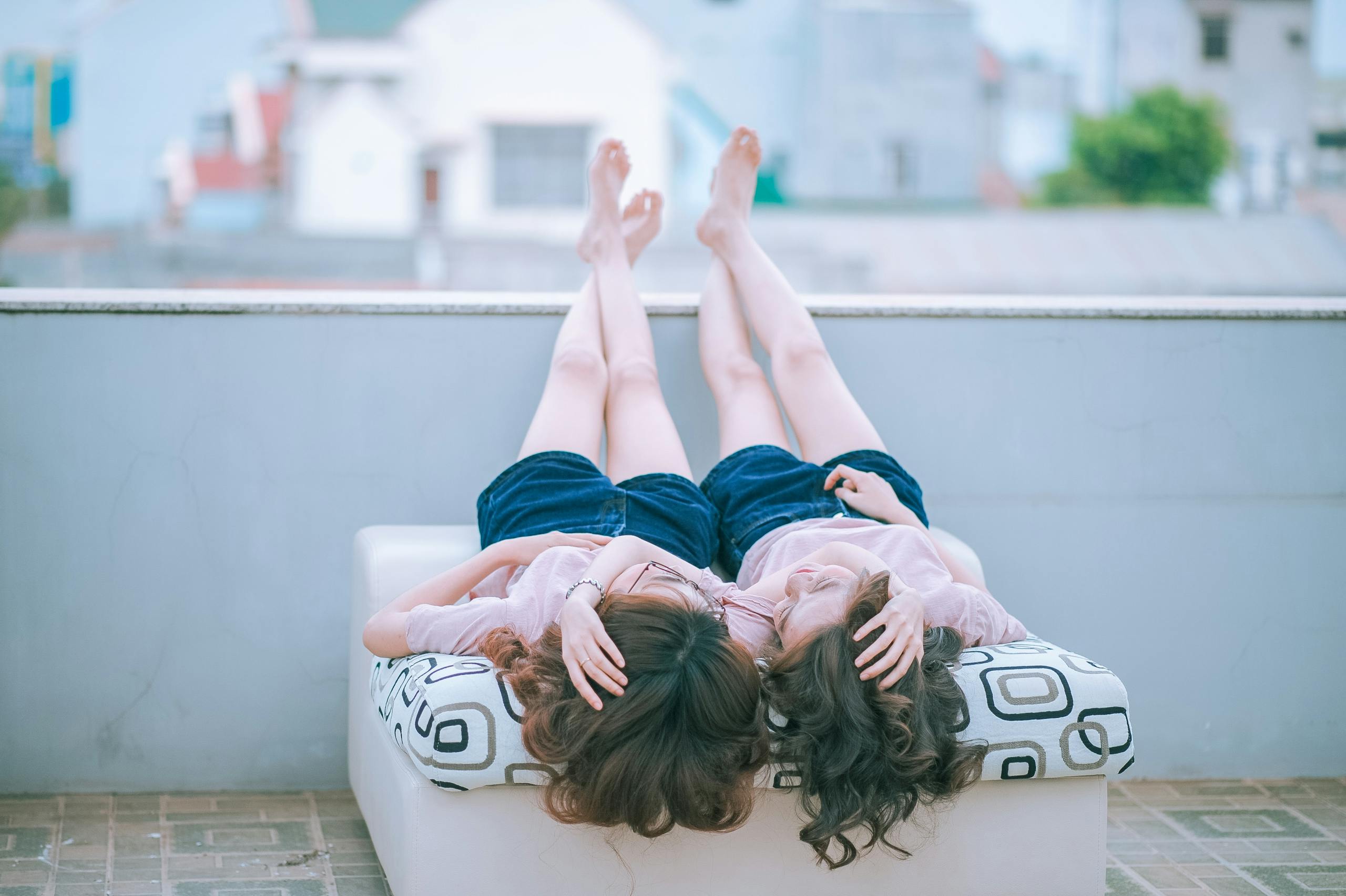 Two Woman Wearing Pink Shirt and Blue Shorts Lying on Sofa Facing Each Other While Both Feet Crossed and Placed on Concrete Wall